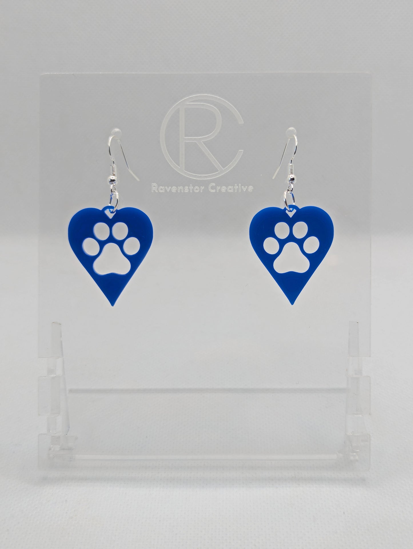 Shows a pair of earrings that have Silver ear wire hook fixings. They are hearts with a dog paw cut out of the middle, perfect for the puppy lover. The colour of them is Blue. They are on see through stands. Against a white background.