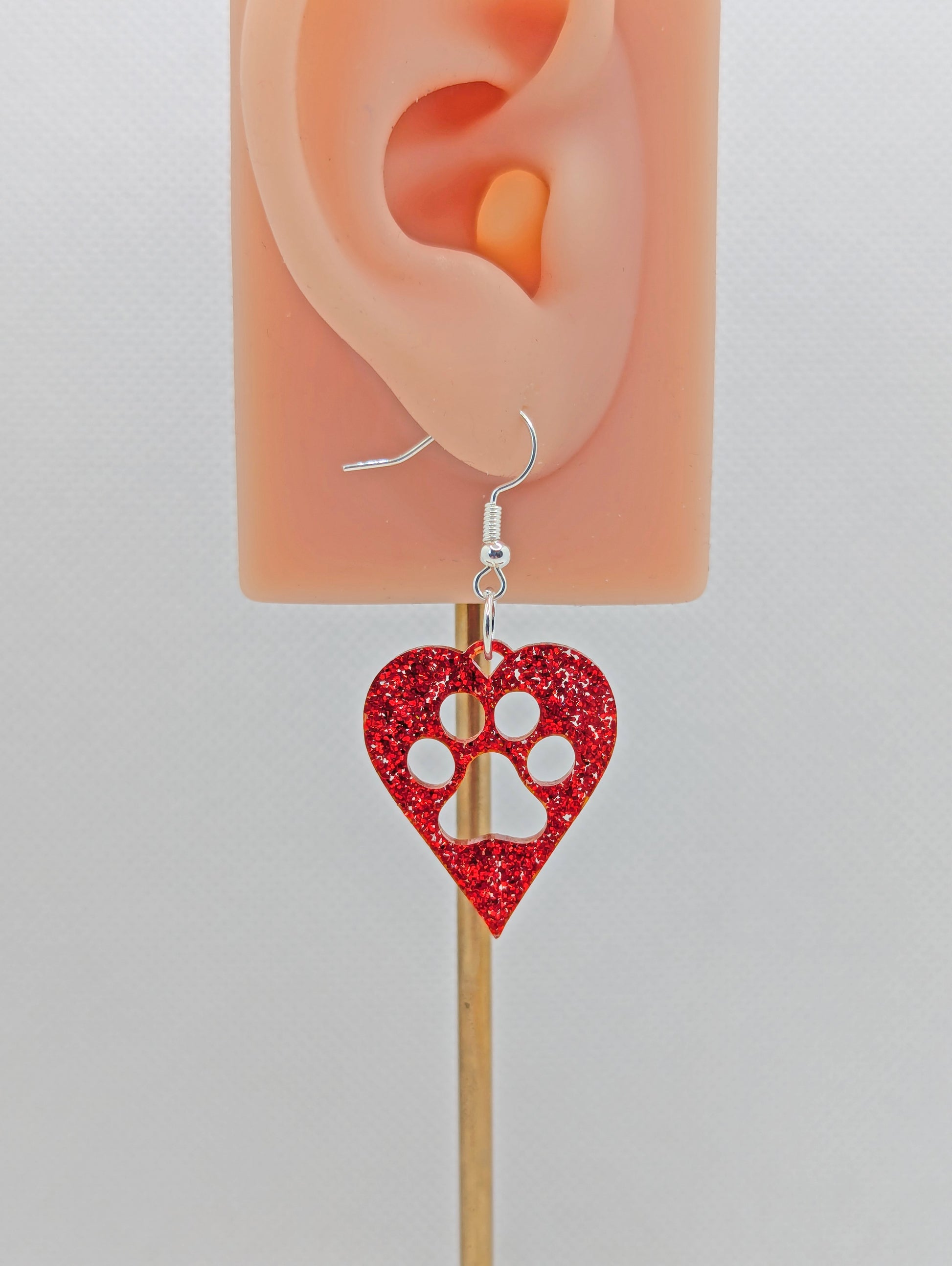 Shows an earring that has a Silver ear wire hook fixing. It is a hearts with a dog paw cut out of the middle, perfect for the puppy lover. The colour of it is Red Glitter. It is on an ear to show the size against a white background.