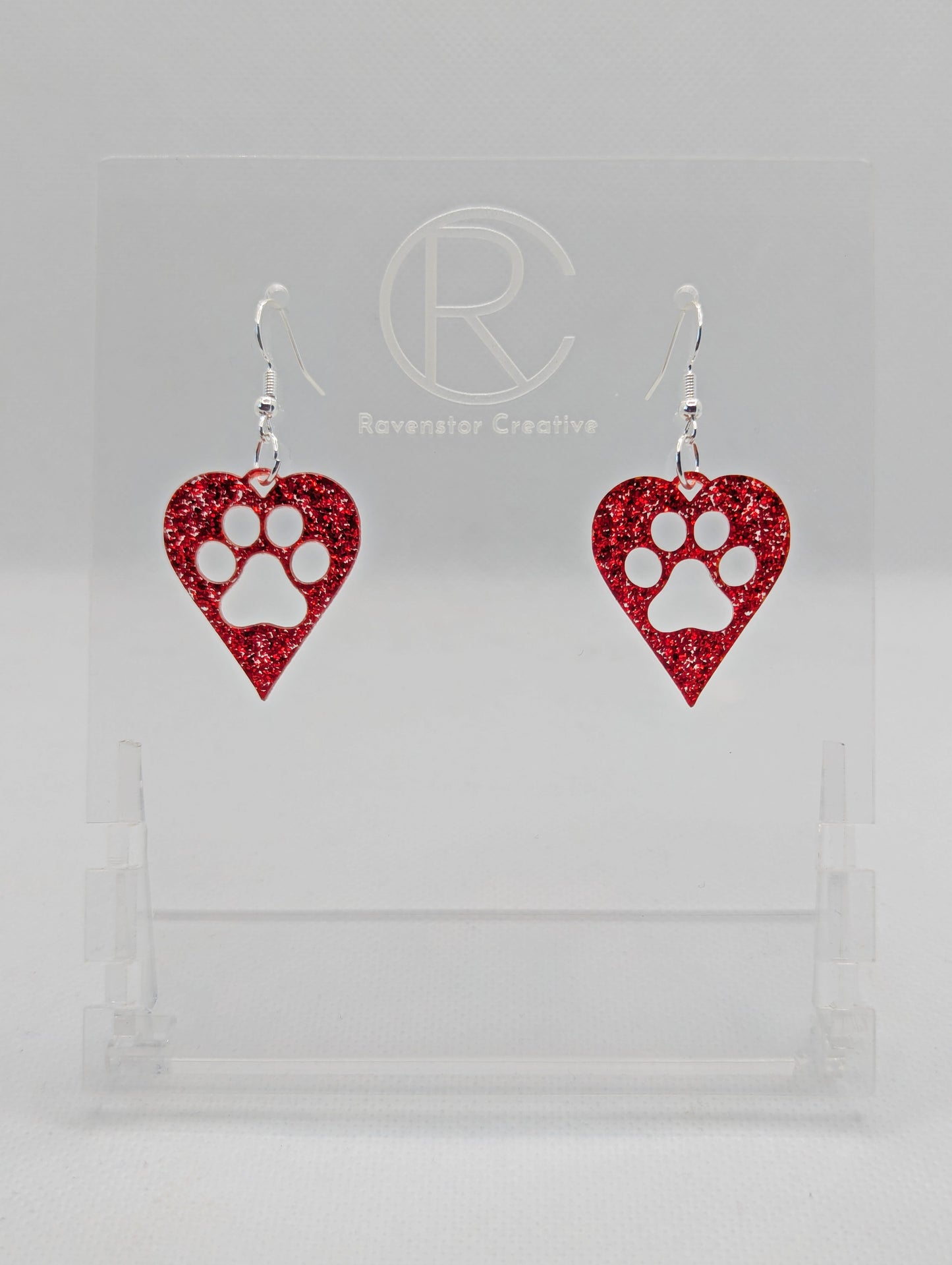 Shows a pair of earrings that have Silver ear wire hook fixings. They are hearts with a dog paw cut out of the middle, perfect for the puppy lover. The colour of them is Red Glitter. They are on see through stands. Against a white background.