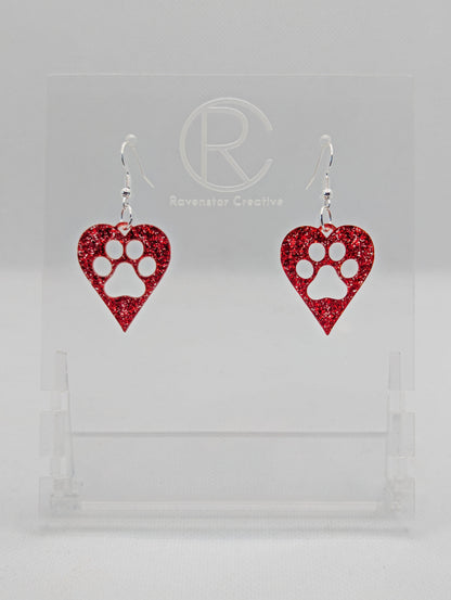 Shows a pair of earrings that have Silver ear wire hook fixings. They are hearts with a dog paw cut out of the middle, perfect for the puppy lover. The colour of them is Red Glitter. They are on see through stands. Against a white background.