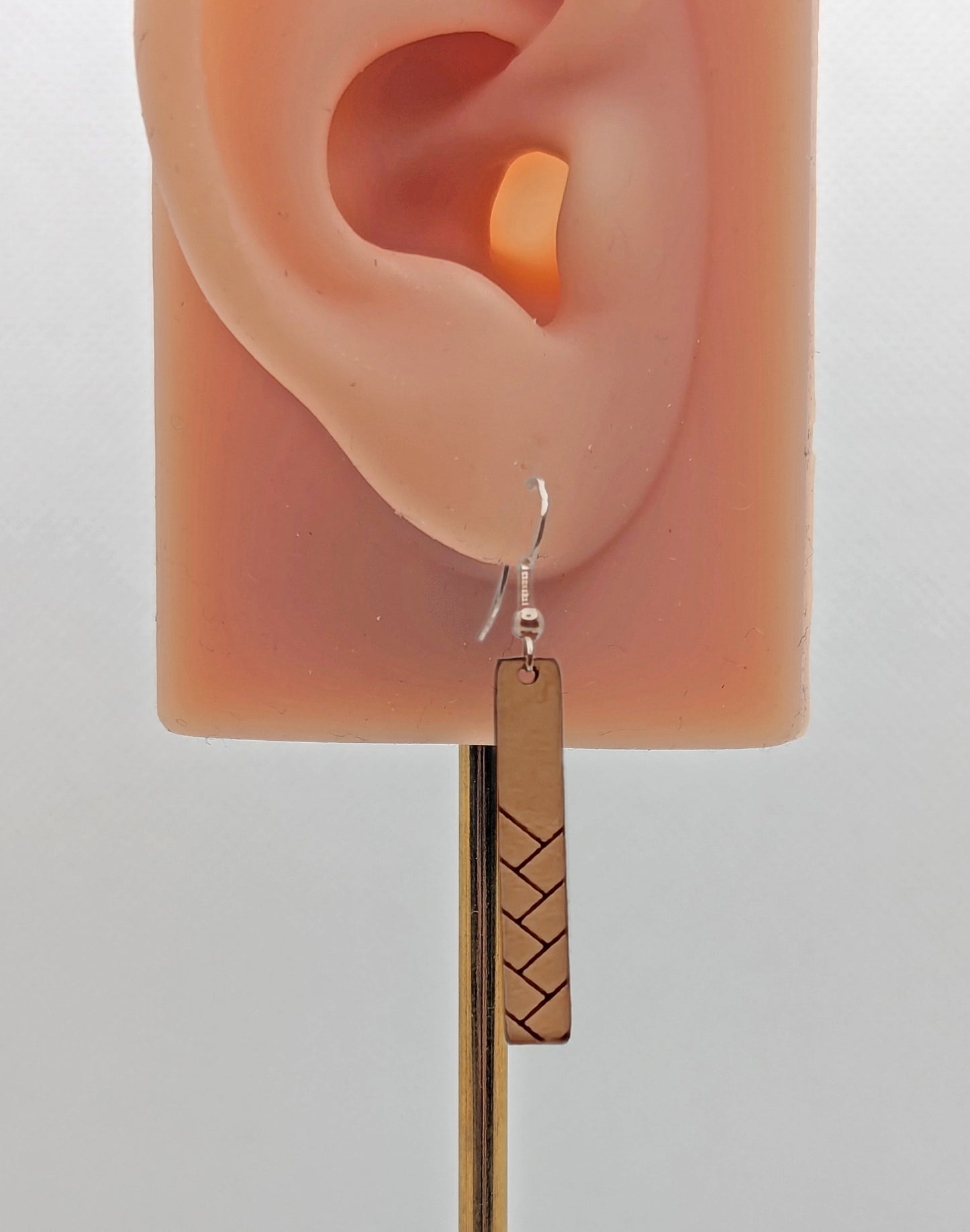 A Natural Tan Leather earring with rose gold fixings. It is thin and long and has a weave pattern. It is on an ear showing the size and is against a white background.