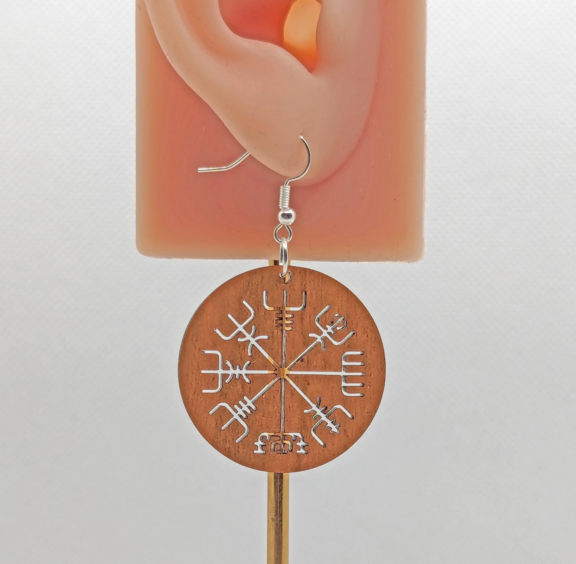 A circle earring with silver ear wire hook fixings. Cut out of the centre is the Nordic Compass charm. It is made from cherry wood. They are on an ear to show the size against a white background.