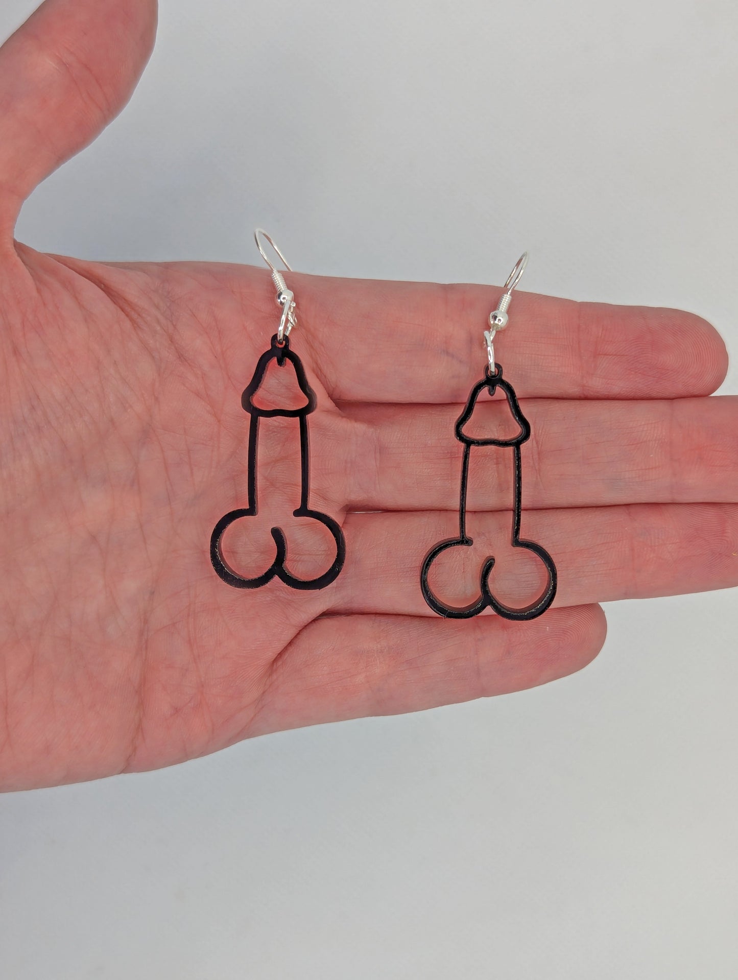 Image of a pair of earrings shaped like a Penis and Balls with Silver ear wire hook fixings. These are ideal for funny Hen Dos or Bachelorette parties. They are against a hand to show the size of two and a half finger widths in length and one wide