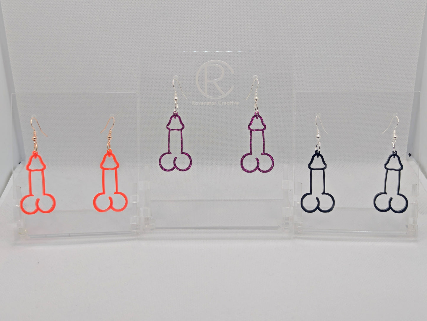 Image of three pairs of Earrings shaped like a Penis and Balls with ear wire hook fixings. These are ideal for funny Hen Dos or  Bachelorette parties. The colours are Black, Pink Glitter and Neon Pink. The are on Clear stands.