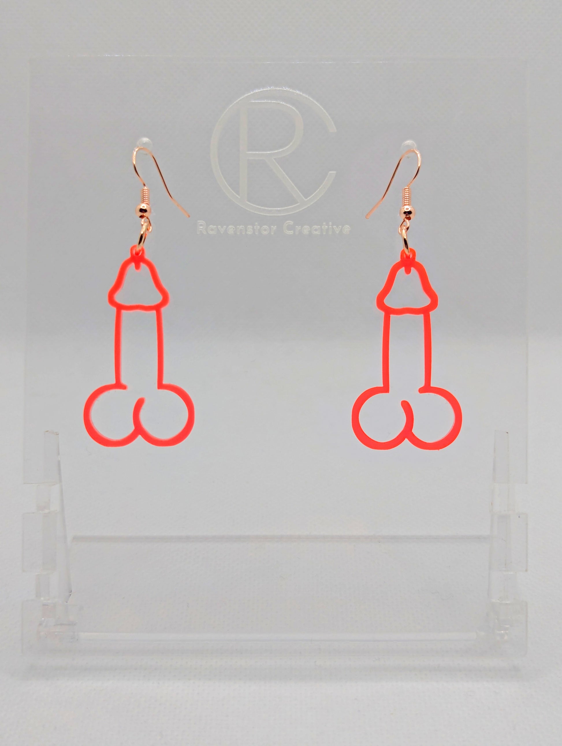 Image of a pair of earrings shaped like a Penis and Balls with Rose Gold ear wire hook fixings. These are ideal for funny Hen Dos or Bachelorette parties. The colour is Neon Pink and they are on clear stands against a white background.