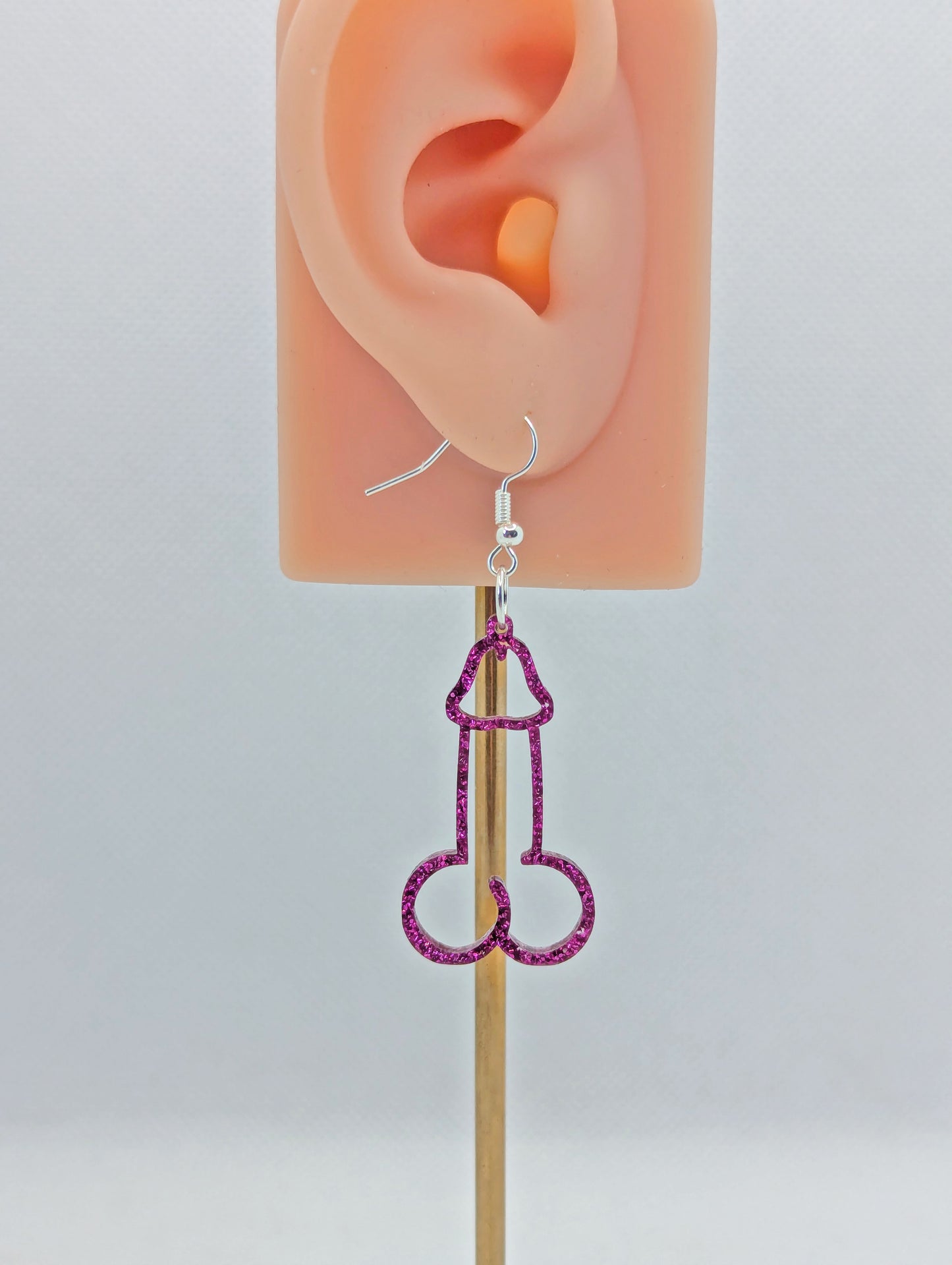 Image of an earring shaped like a Penis and Balls with a Silver ear wire hook fixing. These are ideal for funny Hen Dos or  Bachelorette parties. The colour is Pink Glitter and is on an ear to show the size.