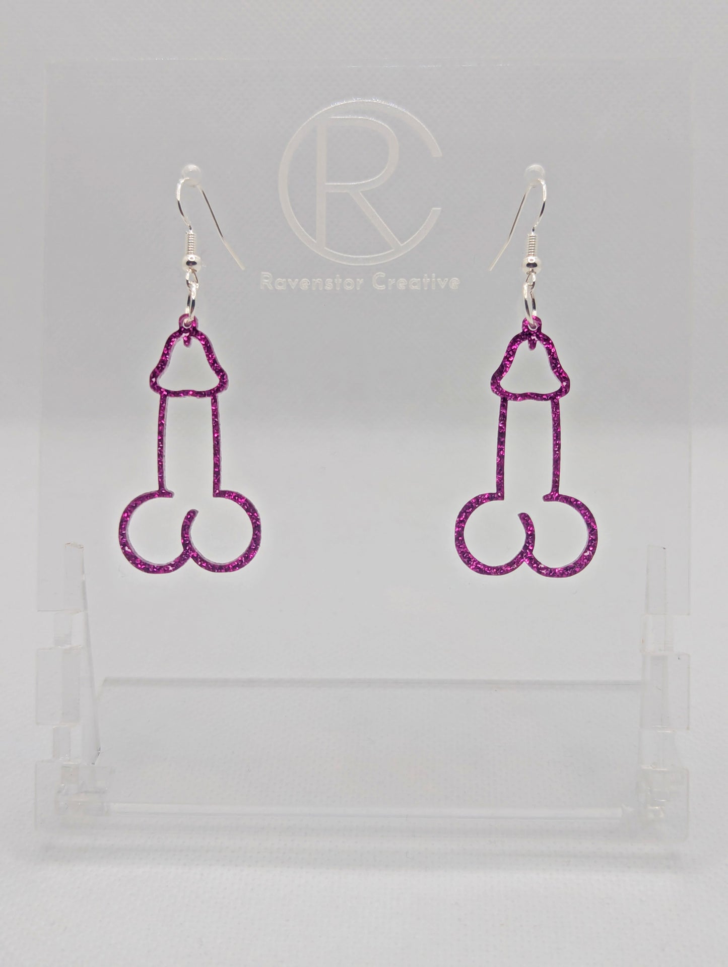 Image of a pair of earrings shaped like a Penis and Balls with Silver ear wire hook fixings. These are ideal for funny Hen Dos or Bachelorette parties. The colour is Pink Glitter and they are on clear stands against a white background.