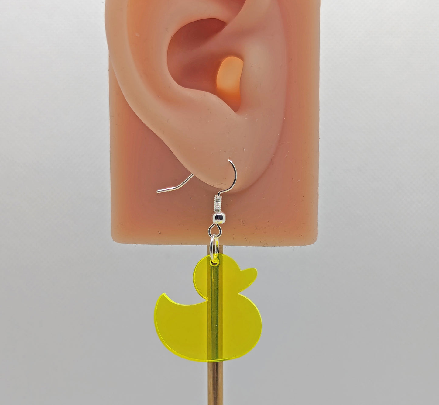 Image of a rubber ducky / duck shaped earring on an ear showing the size against a white background. They are neon yellow and have silver ear wire hook fixings.