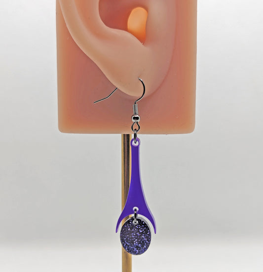 Shows an earring with black ear wire hook fixings. They are slim teardrop shapes in two parts on an ear showing thier size against a white background. The colours are purple top with purple glitter oval.