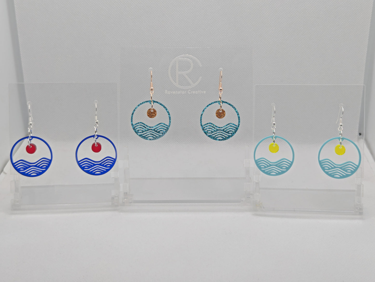 Three pairs of earrings that are two parts. Ear wire fixings are attached to a circle depicting water a small sun dangling in the centre. Colours are Translucent Blue and Red, Ice Blue Glitter and Copper Glitter, Spearmint Blue and Neon Yellow.