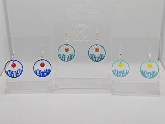 Three pairs of earrings that are two parts. Ear wire fixings are attached to a circle depicting water a small sun dangling in the centre. Colours are Translucent Blue and Red, Ice Blue Glitter and Copper Glitter, Spearmint Blue and Neon Yellow.