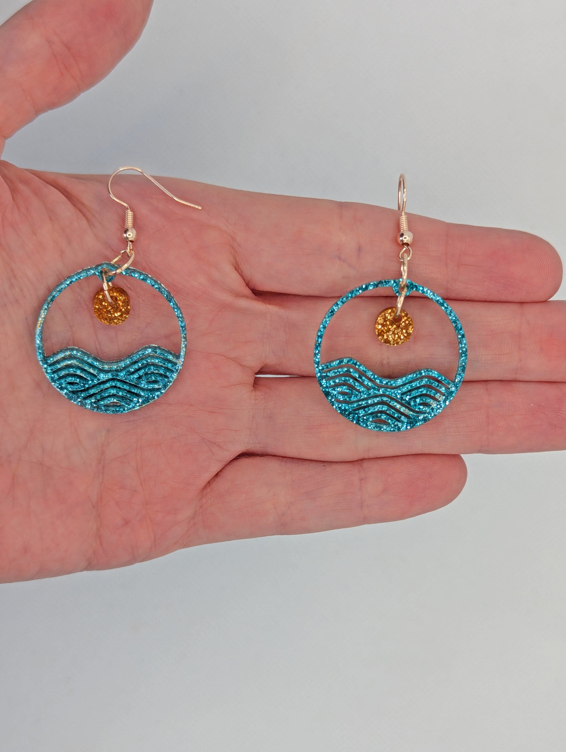 A pair of earrings that are two parts. Silver ear wire fixings are attached to a circle depicting water a small sun dangling in the centre. They are against a hand to show the size of two finger widths wide and long.