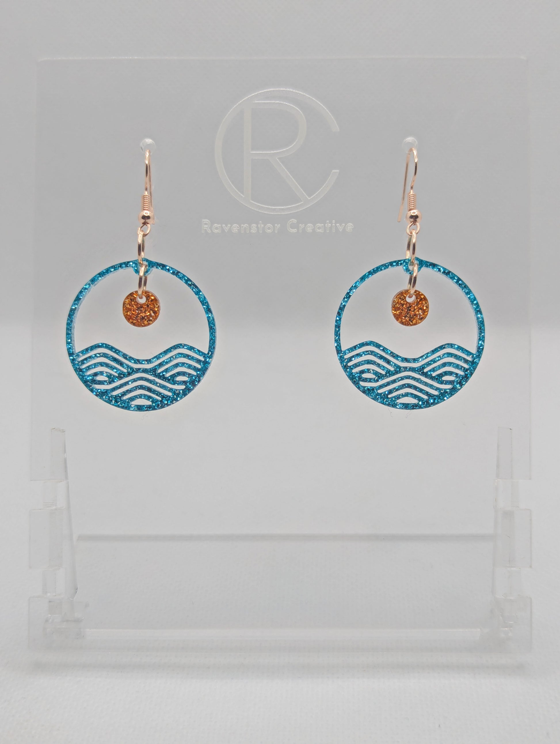 A pair of earrings that are two parts. Rose Gold ear wire fixings are attached to a circle depicting water a small sun dangling in the centre. The colour of the circle is Ice Blue Glitter and the colour of the inner sun is Copper Glitter. They are on a clear stand against a white background.