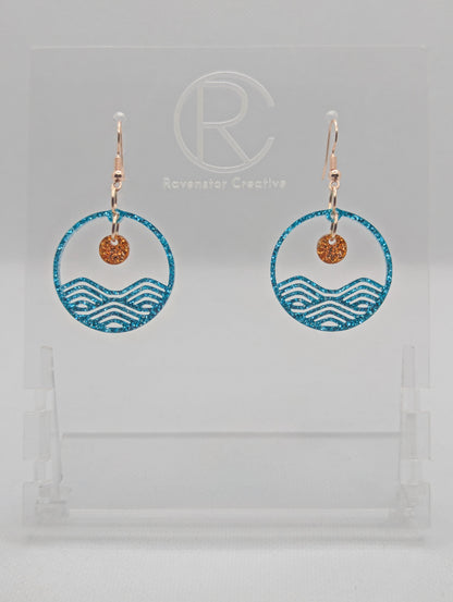 A pair of earrings that are two parts. Rose Gold ear wire fixings are attached to a circle depicting water a small sun dangling in the centre. The colour of the circle is Ice Blue Glitter and the colour of the inner sun is Copper Glitter. They are on a clear stand against a white background.