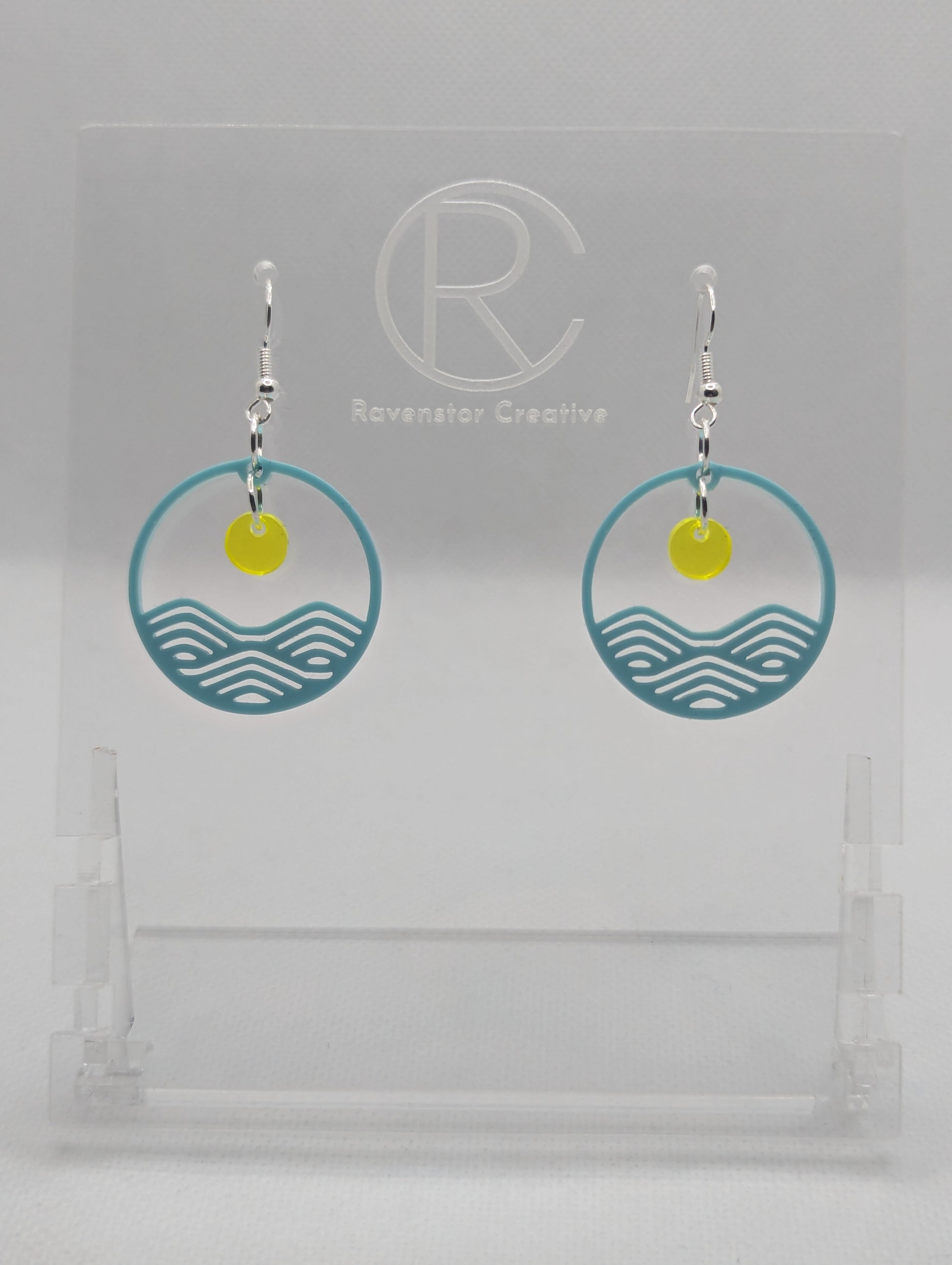 A pair of earrings that are two parts. Silver ear wire fixings are attached to a circle depicting water a small sun dangling in the centre. The colour of the circle is Spearmint and the colour of the inner sun is Neon Yellow. They are on a clear stand against a white background.