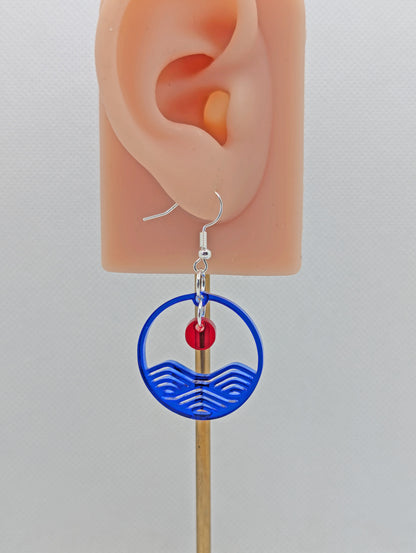 An earring that is two parts. It is a circle depicting water and a small sun dangling in the centre. The colour of the circle is Translucent Blue and the colour of the inner sun is Translucent Red. They are on an ear to show the size.