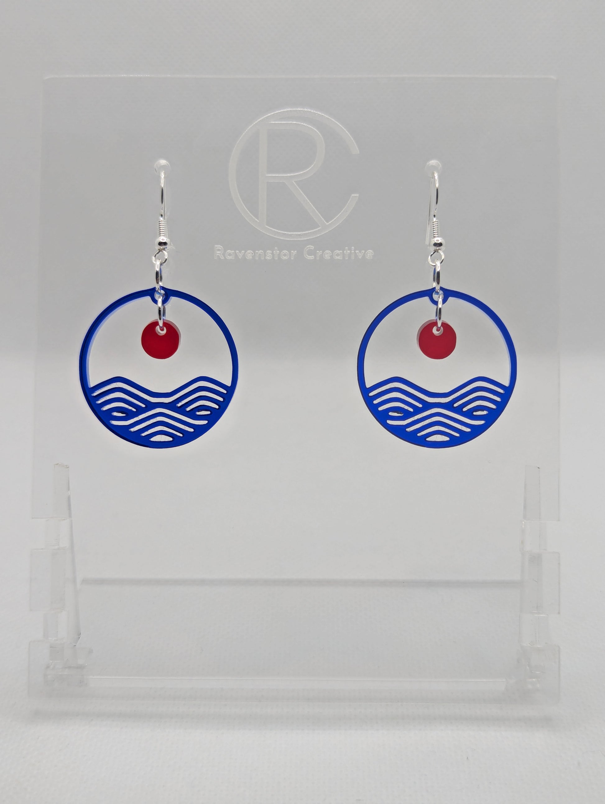 A pair of earrings that are two parts. Silver ear wire fixings are attached to a circle depicting water a small sun dangling in the centre. The colour of the circle is Translucent Blue and the colour of the inner sun is Translucent Red. They are on a clear stand against a white background.