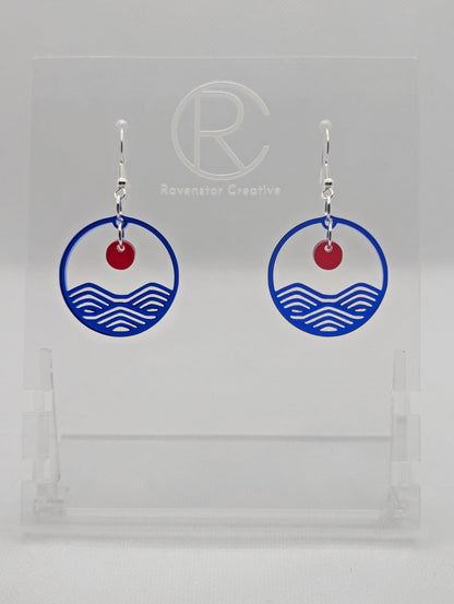 A pair of earrings that are two parts. Silver ear wire fixings are attached to a circle depicting water a small sun dangling in the centre. The colour of the circle is Translucent Blue and the colour of the inner sun is Translucent Red. They are on a clear stand against a white background.