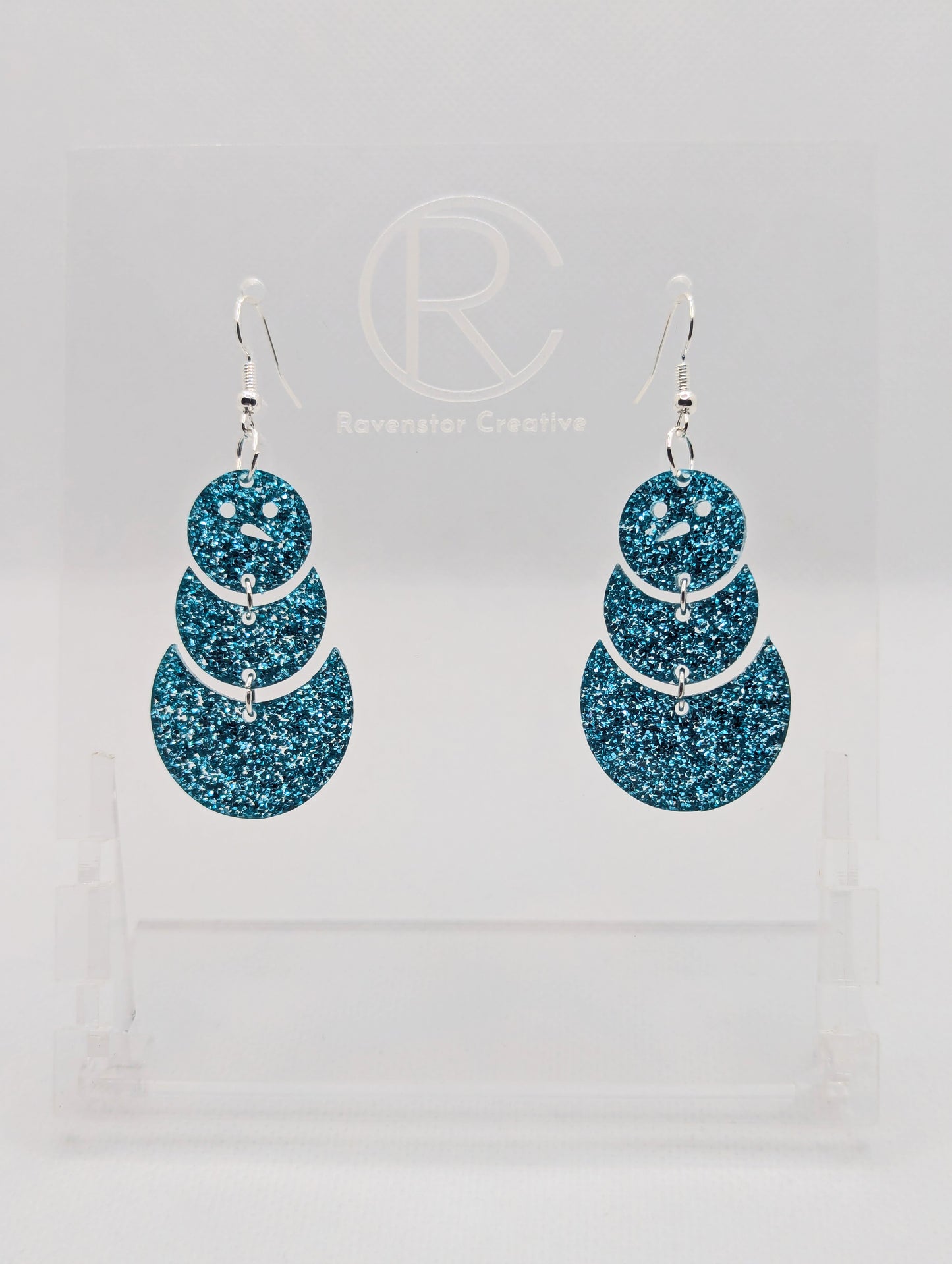 An image of snowman earrings with silver ear wire hook fixings. They are in three parts that dangle. They are made from Ice Blue glitter acrylic and are on a clear stand with the ravenstor creative logo on it against a white background