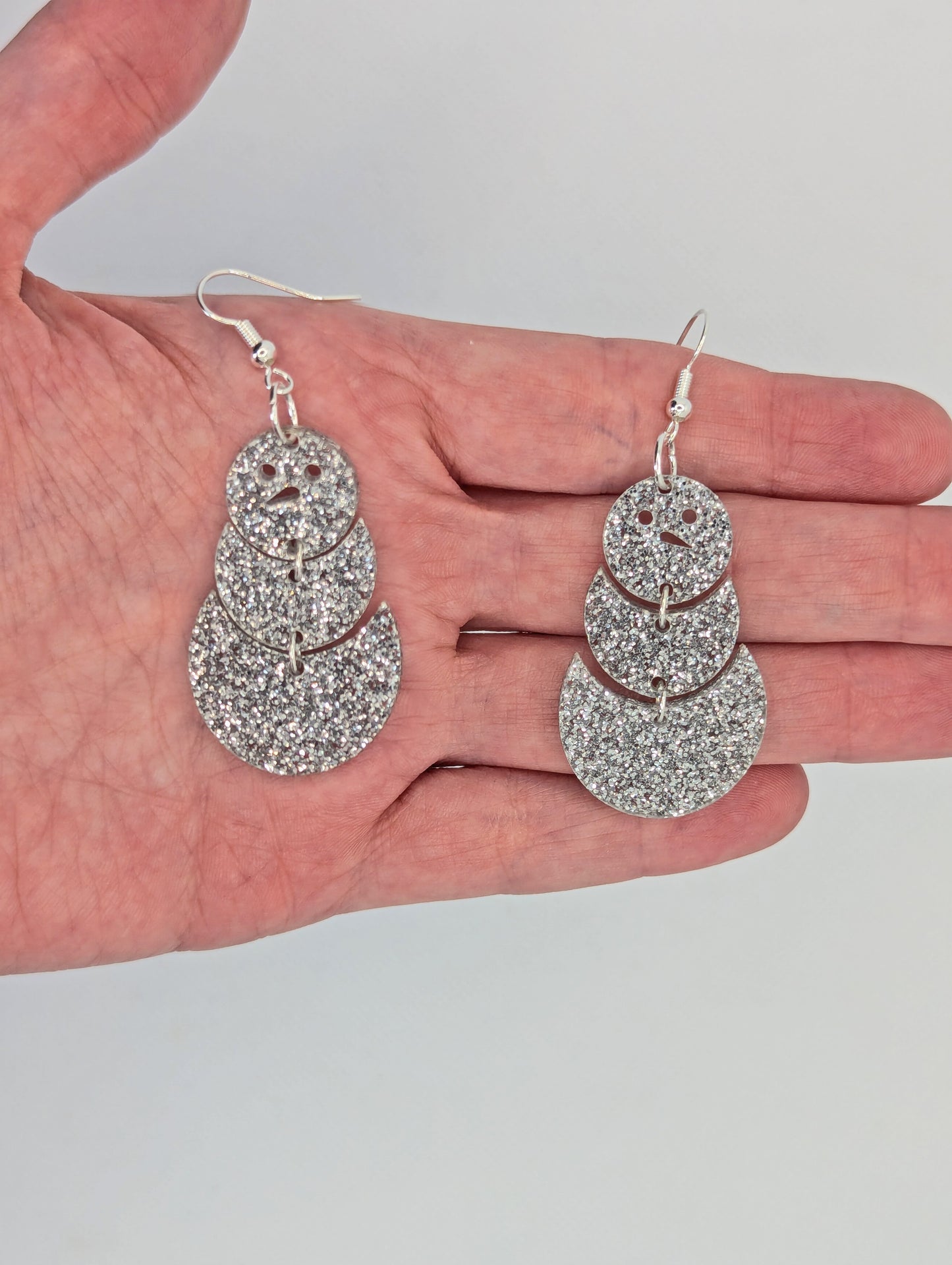 An image of snowman earrings with silver ear wire hook fixings. They are in three parts that dangle. They are made from Silver glitter acrylic and are against a hand to show the size of two and a half finger widths in length and one and a half wide.