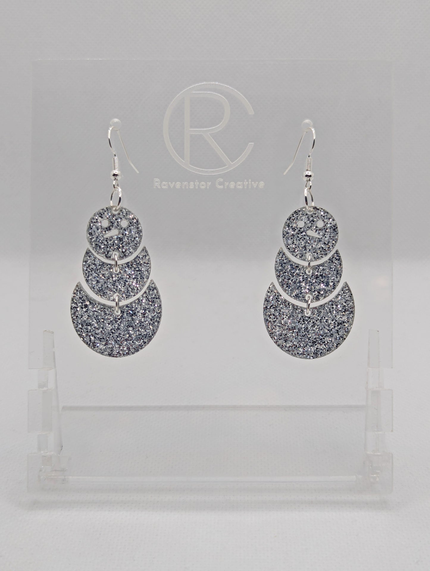 An image of snowman earrings with silver ear wire hook fixings. They are in three parts that dangle. They are made from Silver glitter acrylic and are on a clear stand with the ravenstor creative logo on it against a white background