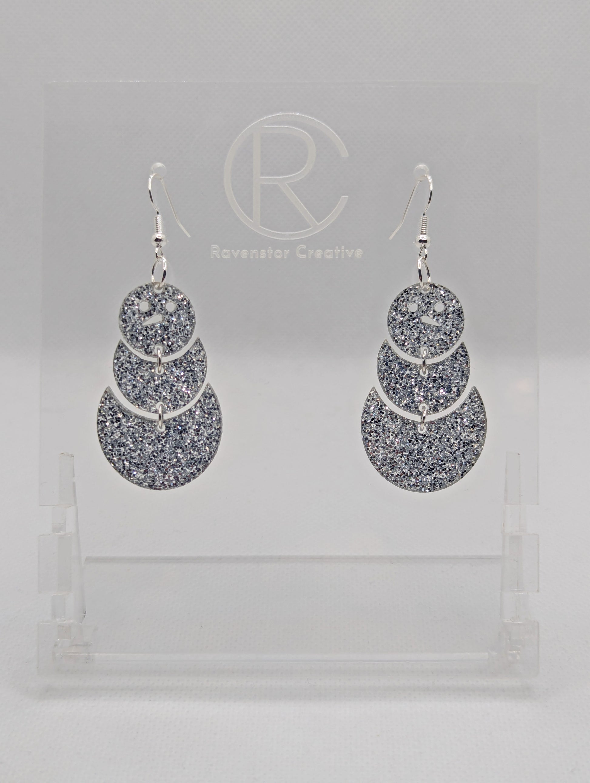 An image of snowman earrings with silver ear wire hook fixings. They are in three parts that dangle. They are made from Silver glitter acrylic and are on a clear stand with the ravenstor creative logo on it against a white background