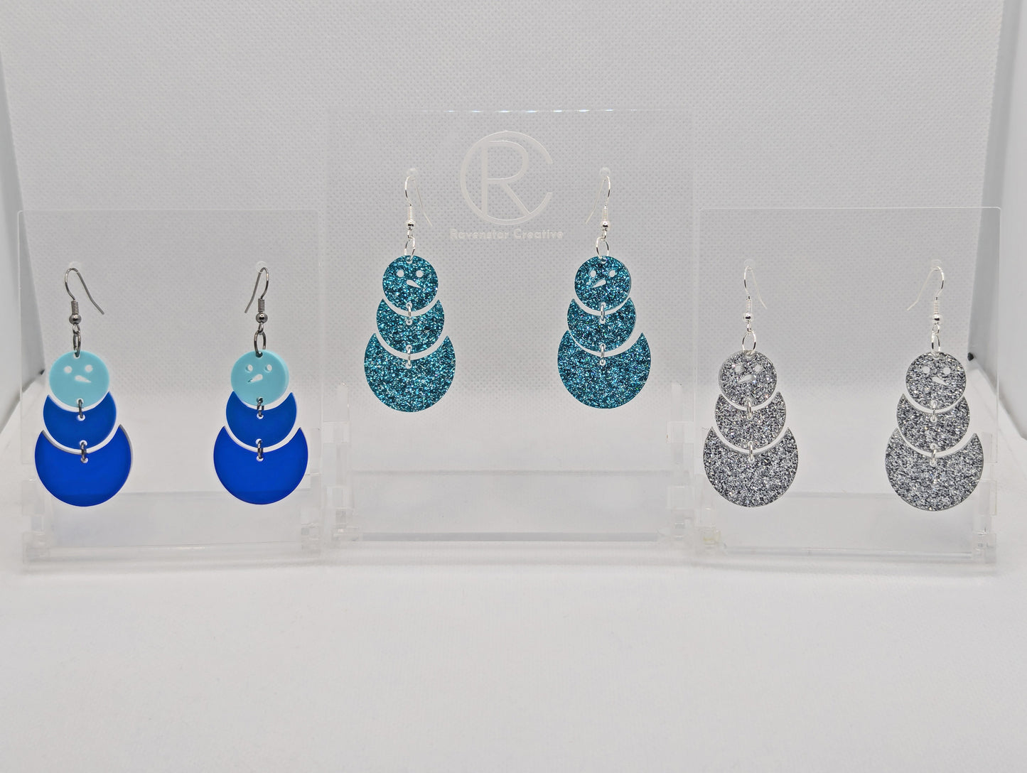 An image of three pairs of Snowman earrings with ear wire hook fixings. They are in three parts that dangle. The are made from acrylic and are Silver glitter, ice blue glitter and Spearmint, blue and Translucent Blue in colour.