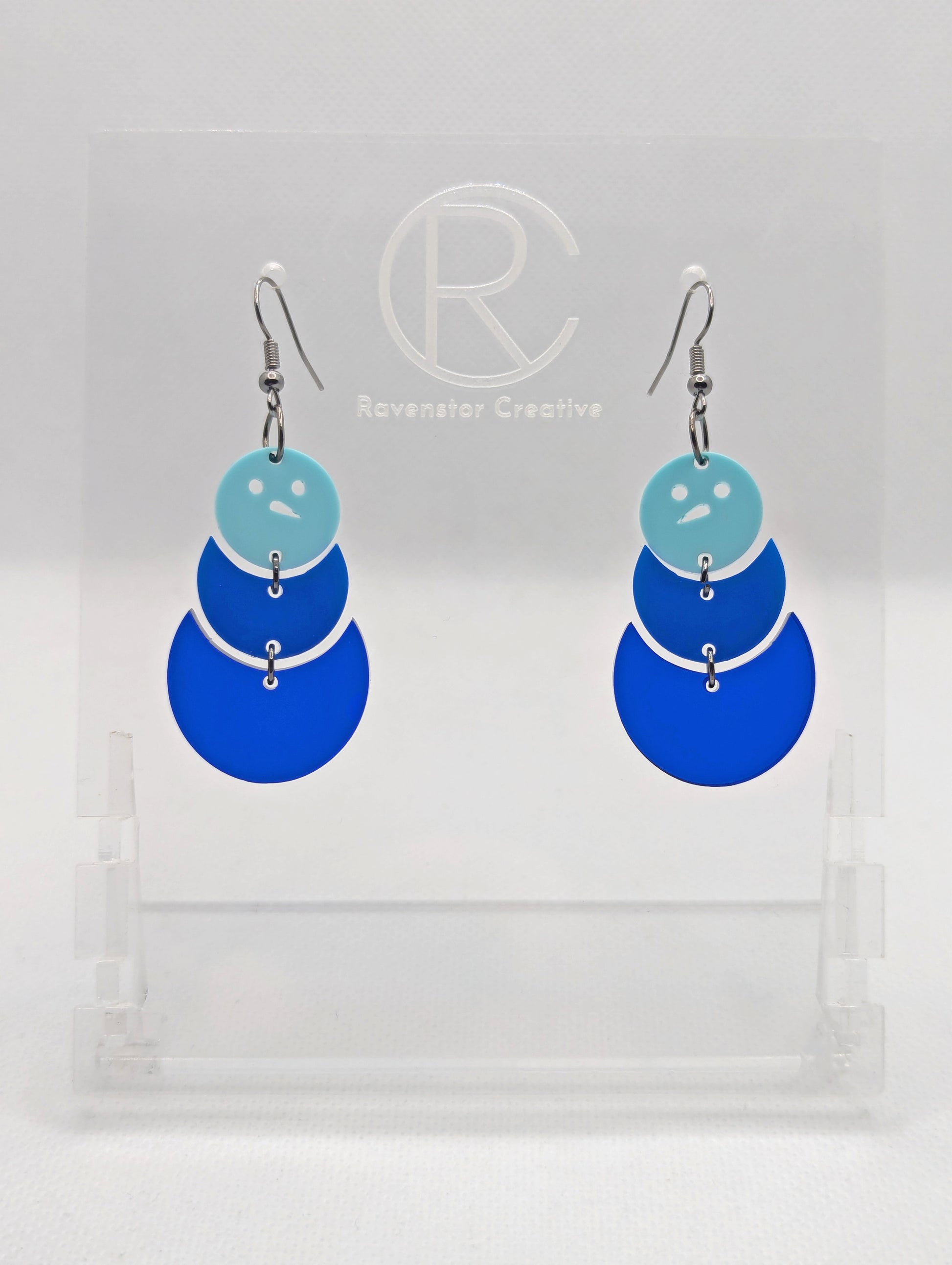 An image of snowman earrings with black ear wire hook fixings. They are in three parts that dangle. They are made from Spearmint, blue and translucent blue acrylic and are on a clear stand against a white background