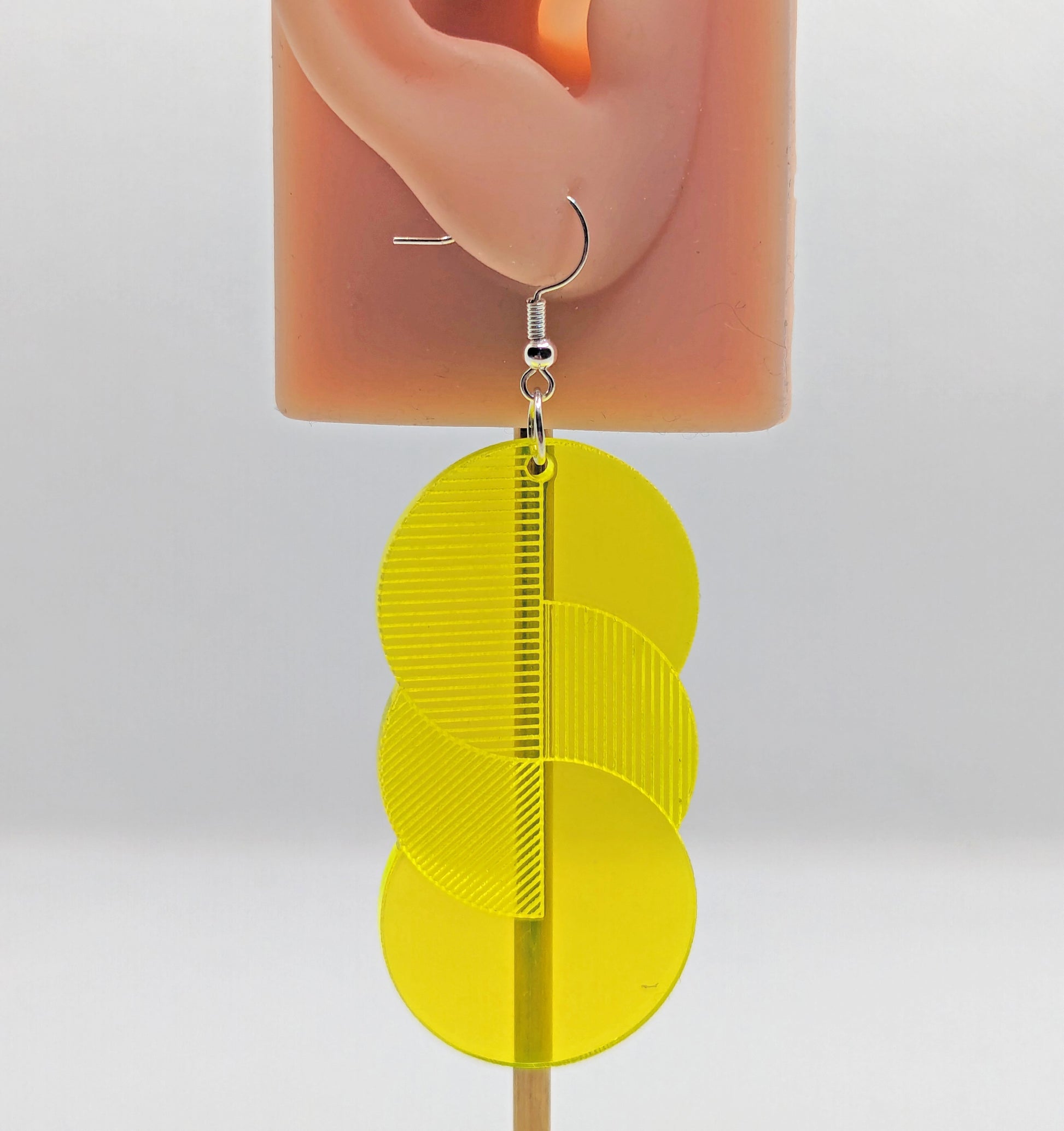 A large acrylic earring with silver ear wire hook fixings on an ear to show the size. It is 3 circles overlapping and entwined with  engraved line decorations. They are made from Neon Orange acrylic.