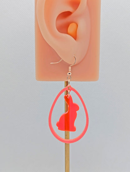 A two part drop earring with a Rose Gold ear wire hook fixing. It is an egg shaped frame with a Bunny Rabbit dangling inside. The colours are Neon Pink for the Bunny and Sherbet Pink for the Egg Frame. It is on an ear to show the size.