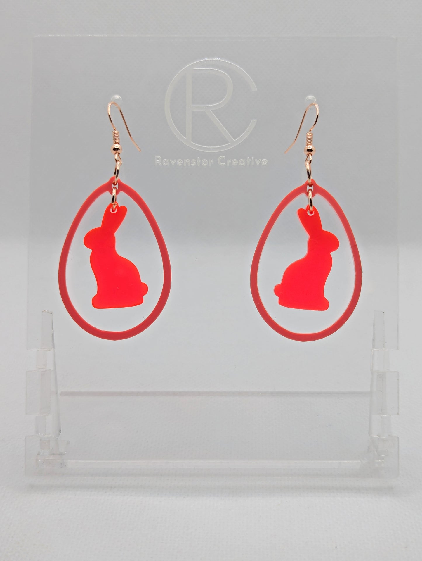 A pair of two part drop earrings with Rose Gold ear wire hook fixings. They are an egg shaped frame with a Bunny Rabbit dangling inside. The colours are Neon Pink for the Bunny and Sherbet Pink for the Egg Frame. The are on a clear stand.