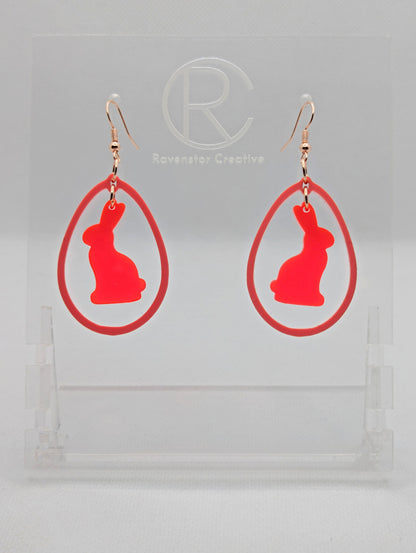 A pair of two part drop earrings with Rose Gold ear wire hook fixings. They are an egg shaped frame with a Bunny Rabbit dangling inside. The colours are Neon Pink for the Bunny and Sherbet Pink for the Egg Frame. The are on a clear stand.