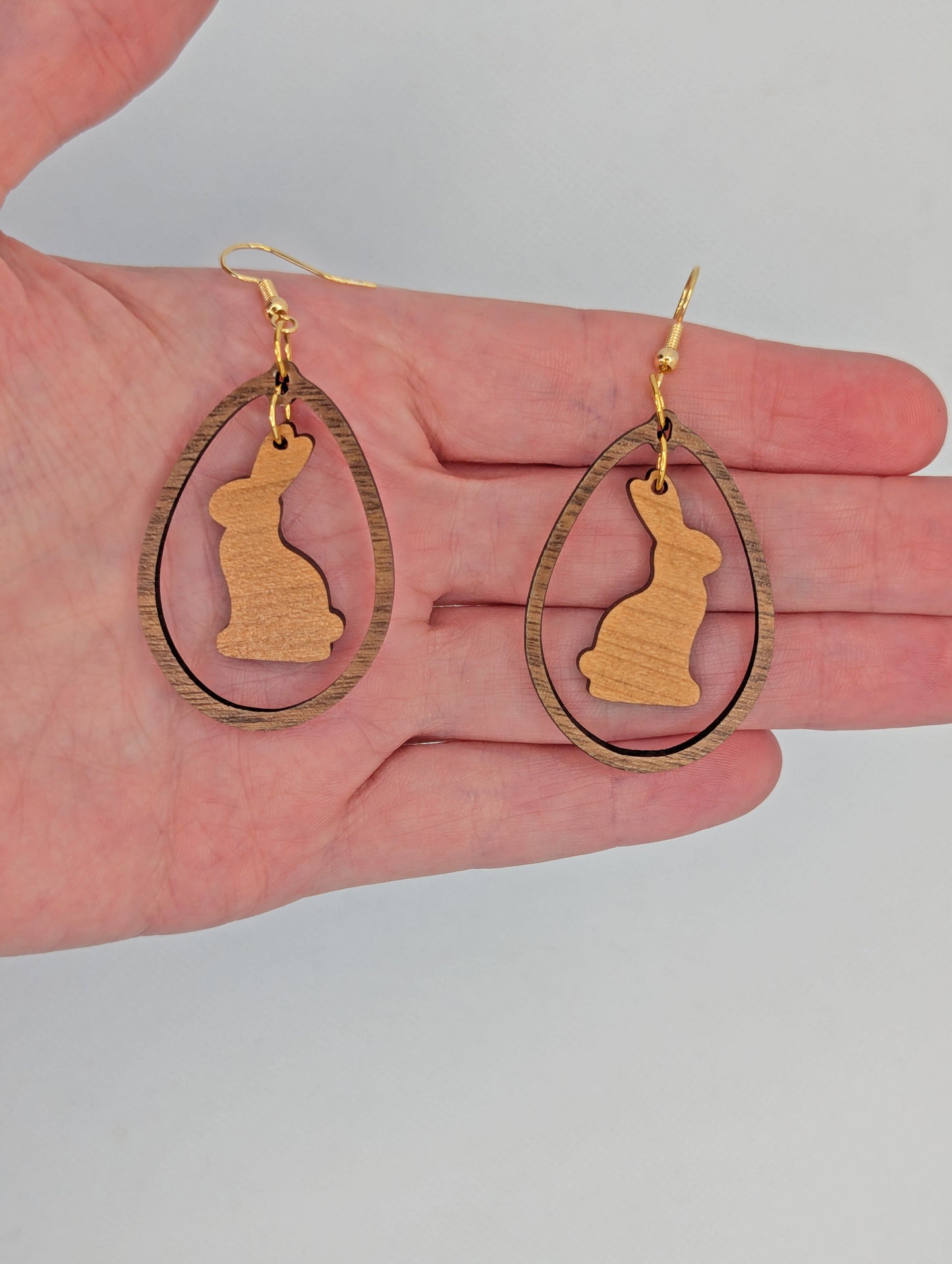 A pair of two part drop earrings with Gold ear wire hook fixings. They are an egg shaped frame with a Bunny Rabbit dangling inside. They are against a hand to show the size of Three Finger widths in length and two wide.