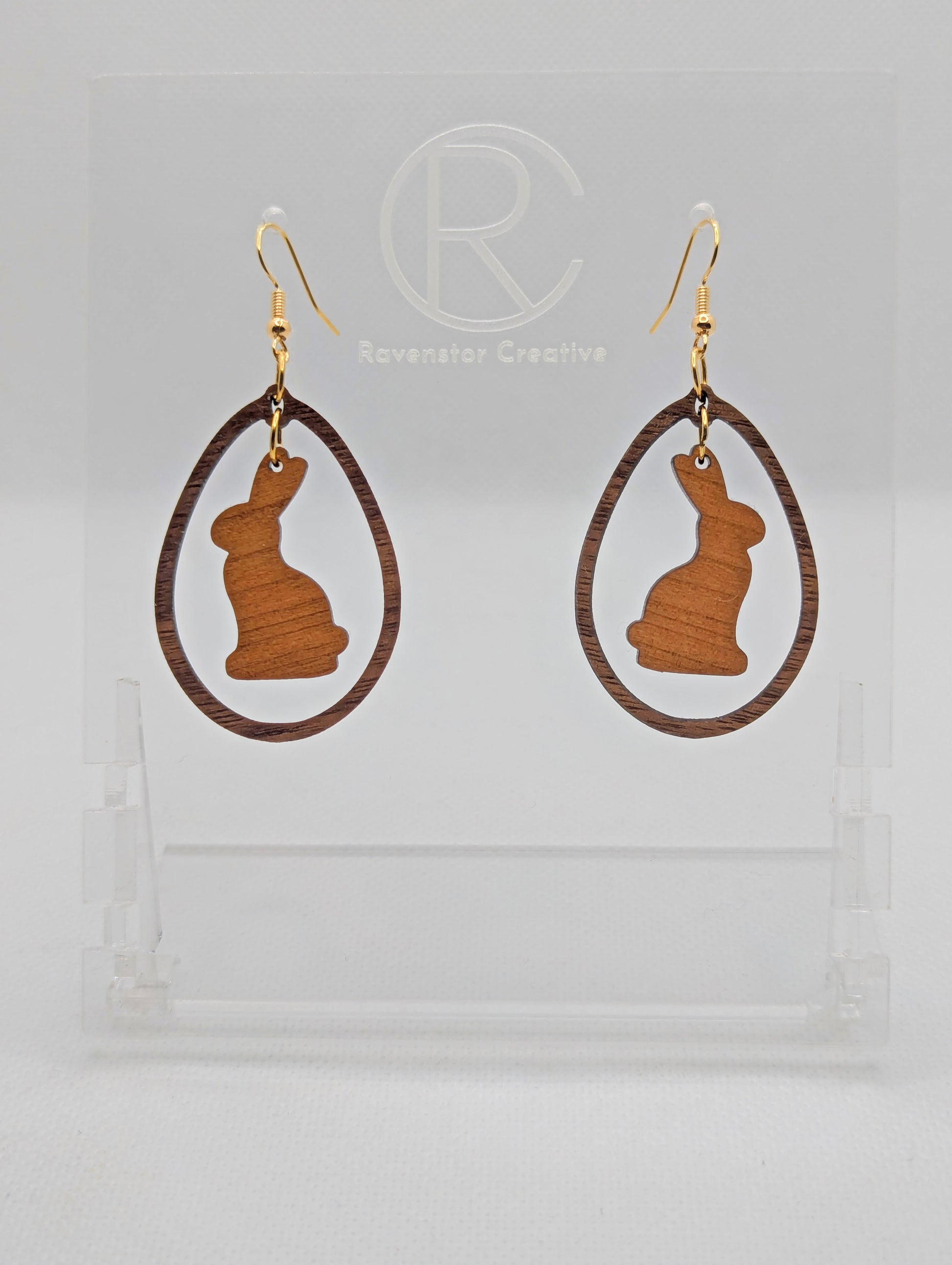 A pair of two part drop earrings with Gold ear wire hook fixings. They are an egg shaped frame with a Bunny Rabbit dangling inside. The colours are Cherry Wood for the Bunny and Walnut Wood for the Egg Frame. The are on a clear stand.