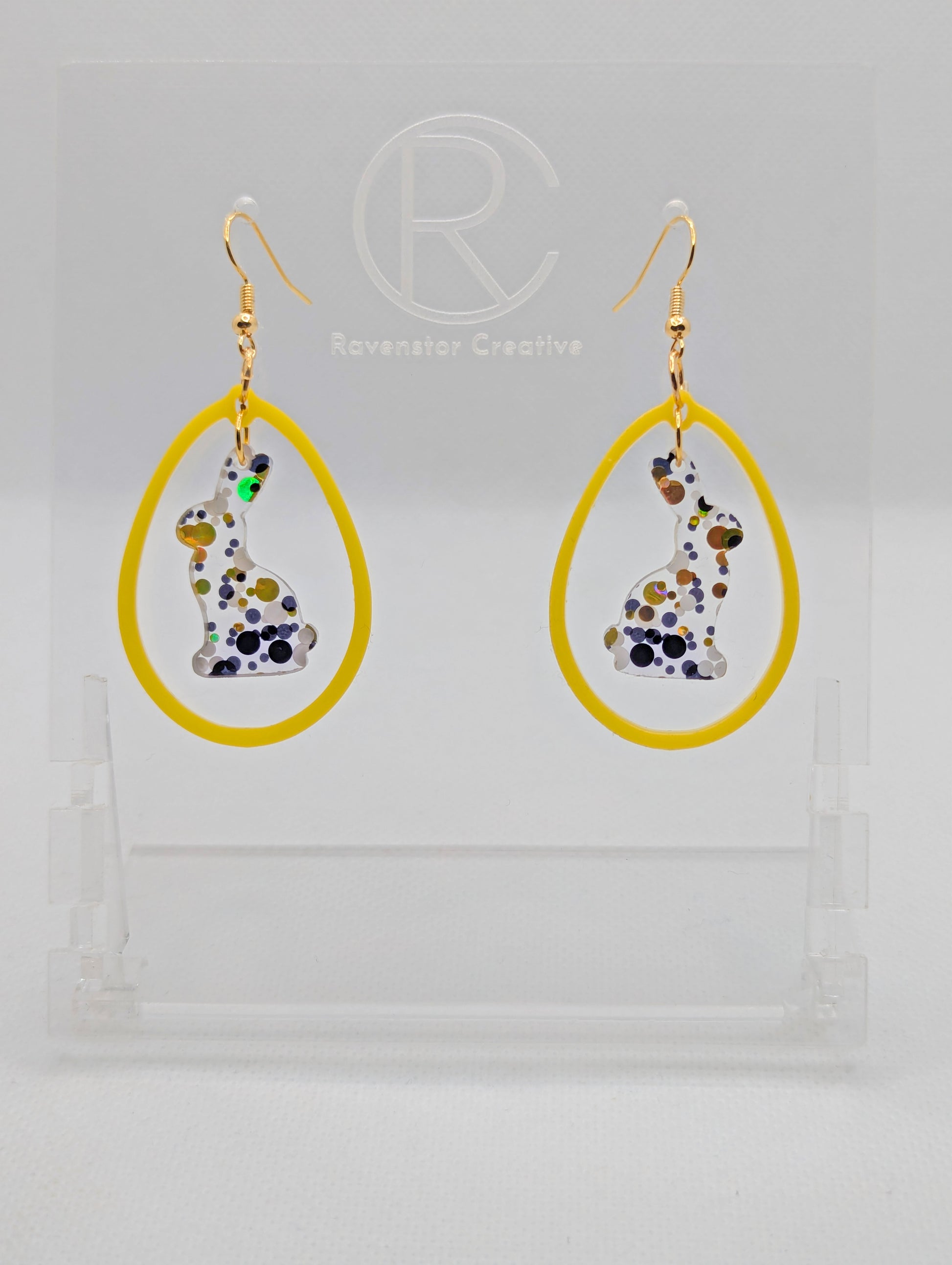 A pair of two part drop earrings with Gold ear wire hook fixings. They are an egg shaped frame with a Bunny Rabbit dangling inside. The colours are Lollipop Glitter for the Bunny and Yellow for the Egg Frame. The are on a clear stand.