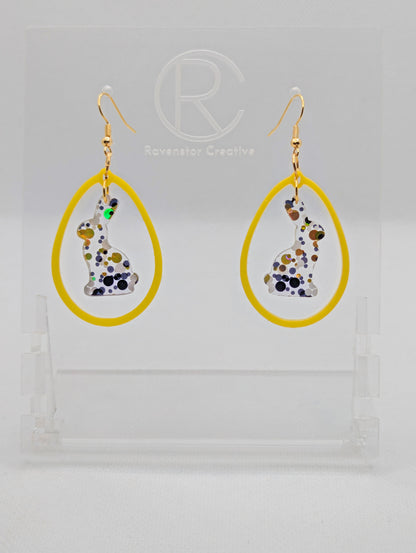 A pair of two part drop earrings with Gold ear wire hook fixings. They are an egg shaped frame with a Bunny Rabbit dangling inside. The colours are Lollipop Glitter for the Bunny and Yellow for the Egg Frame. The are on a clear stand.