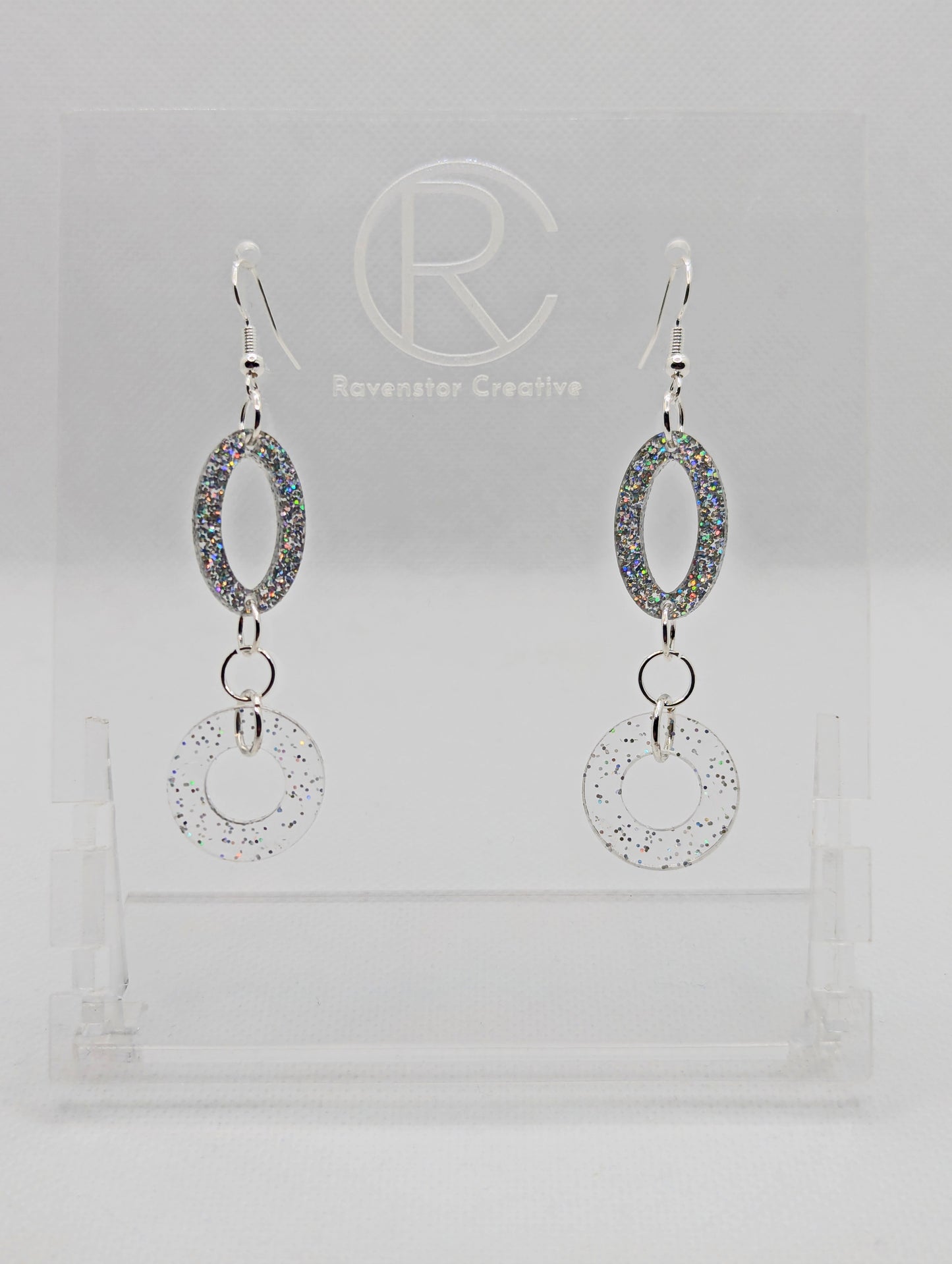 A pair of two part drop earrings with Silver ear wire hook fixings. They are an oval with a circle dangling underneath joined by three Silver jump rings. The colours are Holographic Glitter for the oval and Clear sparkle for the circle.