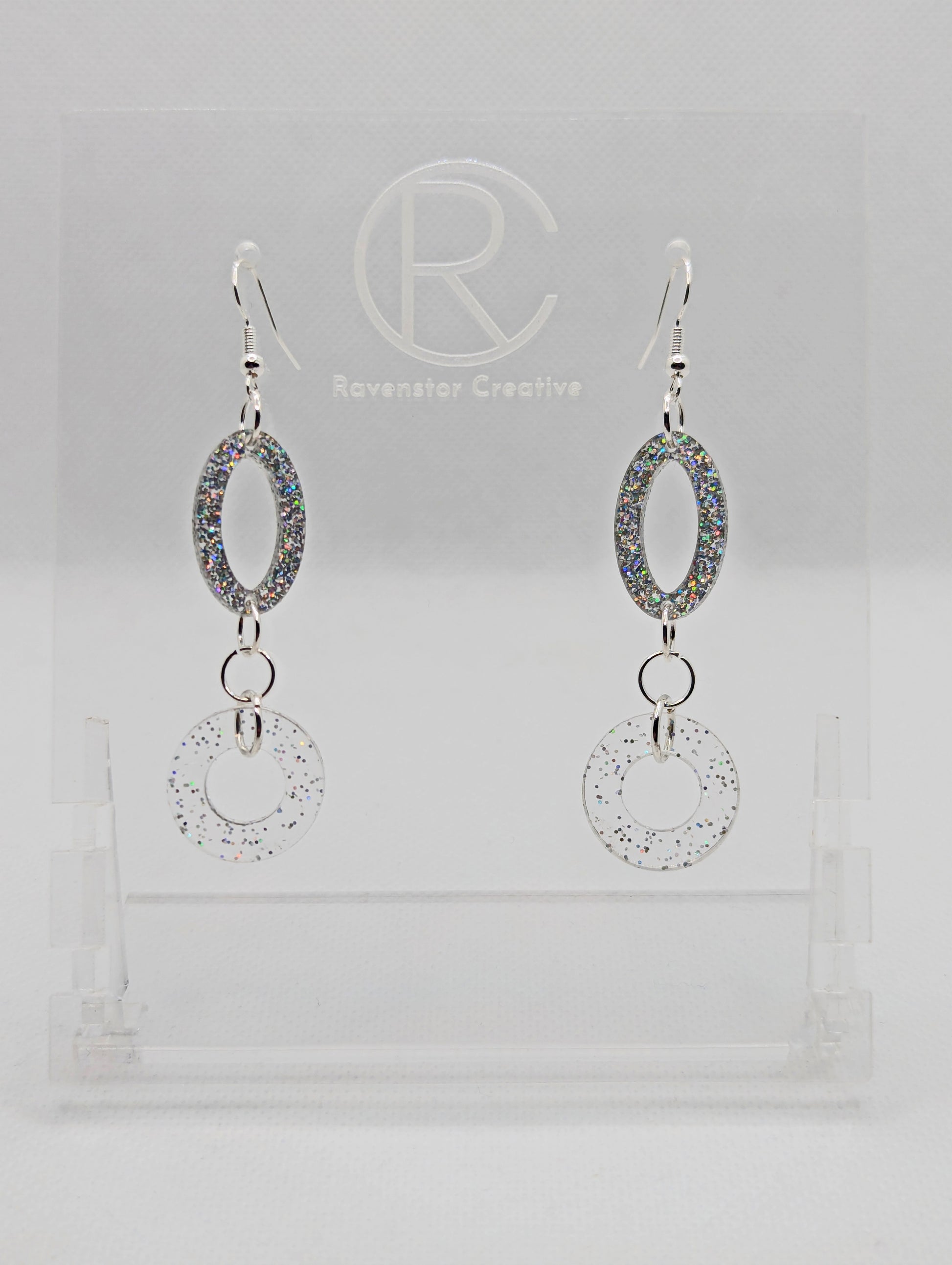 A pair of two part drop earrings with Silver ear wire hook fixings. They are an oval with a circle dangling underneath joined by three Silver jump rings. The colours are Holographic Glitter for the oval and Clear sparkle for the circle.