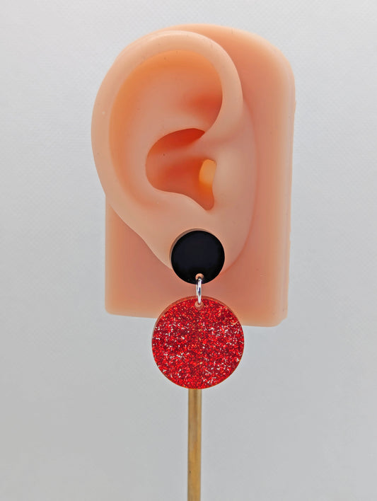 An image of a pair of stud earrings, They are two circles with a smaller one at the top with the stud fixing and a larger one dangling underneath. The colours are Black for the top with a Red Glitter larger circle. It is on an ear to show the size.