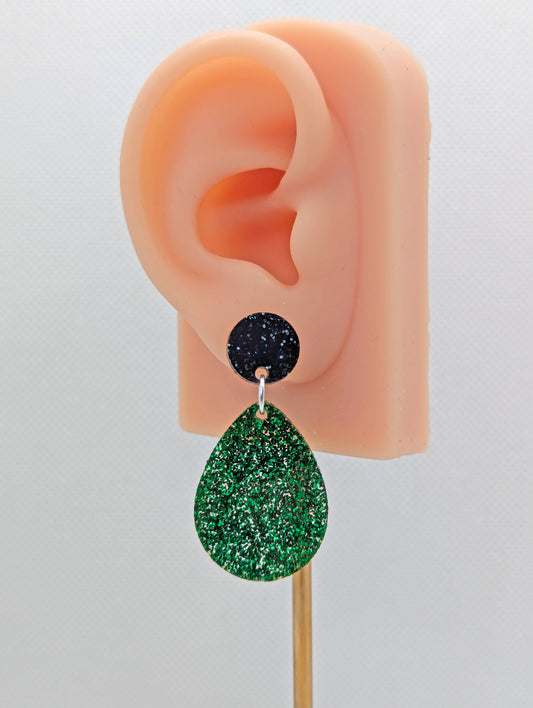 An image of a pair of stud earrings. They are a circle at the top and a larger teardrop shape dangling underneath. The Colours are Black and silver glitter for the circle and Green Glitter for the teardrop. It is on an ear to show the size.