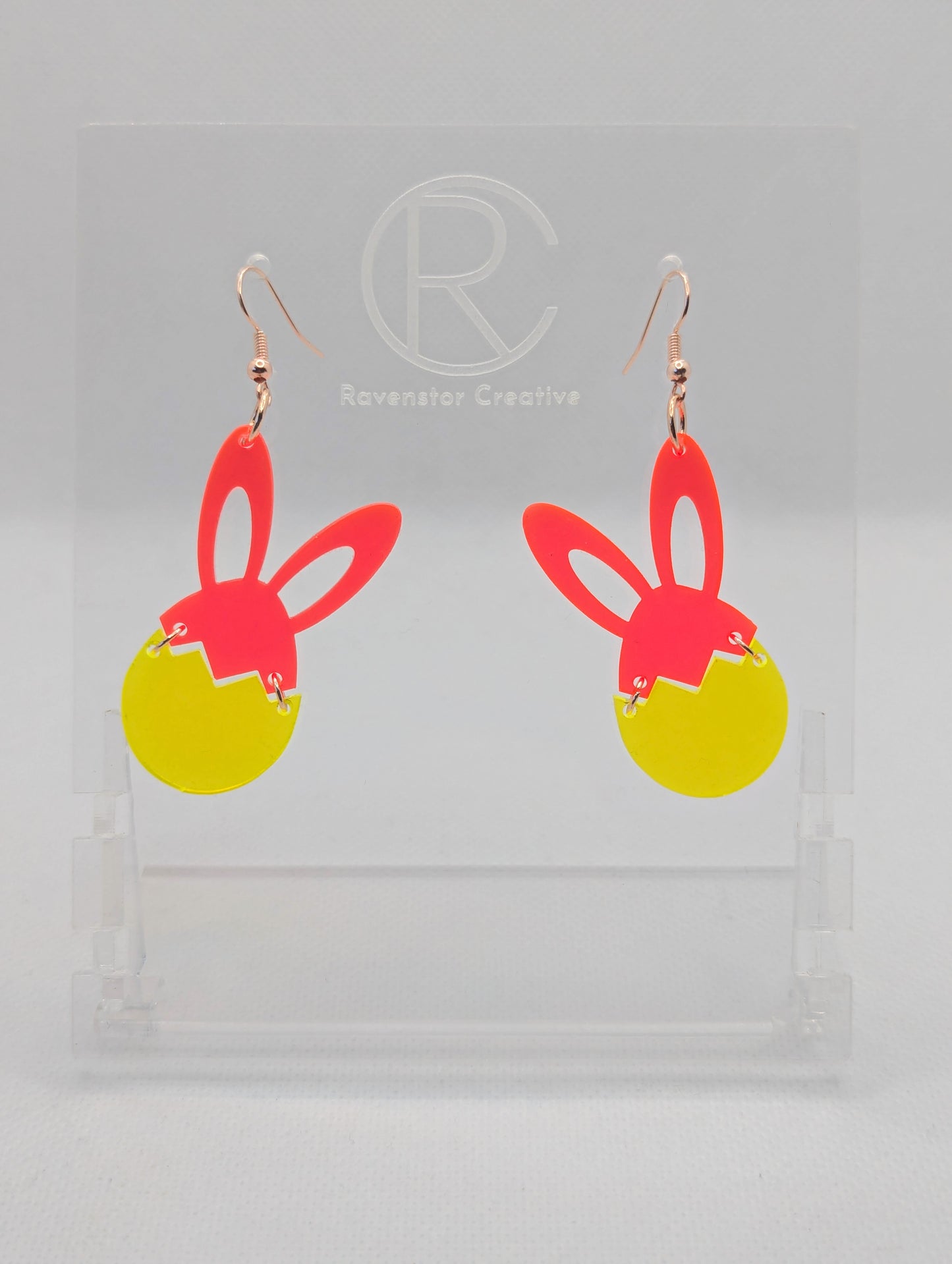 Two part drop earrings with rose gold ear wire hook fixings. The top part are bunny ears and the bottom is a broken egg joined by jump rings. The colours are Neon Pink for the ears and Neon Orange for the egg. They are in a clear stand.