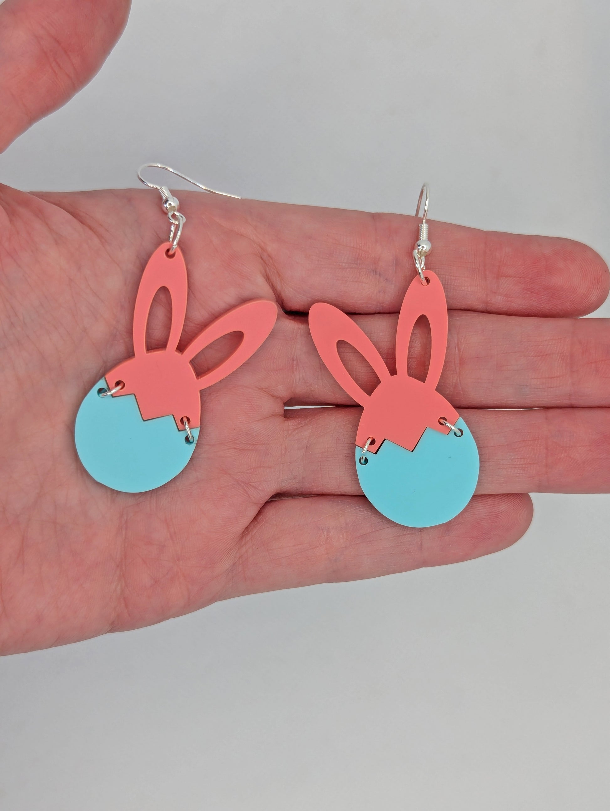 Two part drop earrings with silver ear wire hook fixings. The top part are bunny ears and the bottom is a broken egg joined by jump rings. They are against a hand to show the size of three finger widths in length and two wide.