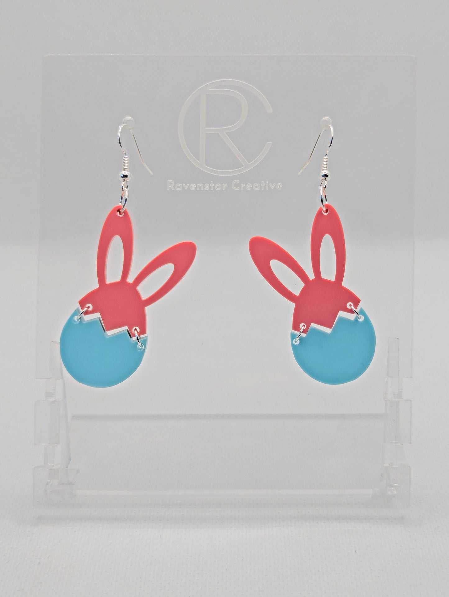 Two part drop earrings with silver ear wire hook fixings. The top part are bunny ears and the bottom is a broken egg joined by jump rings. The colours are Sherbet Pink for the ears and Spearmint for the egg. They are in a clear stand.