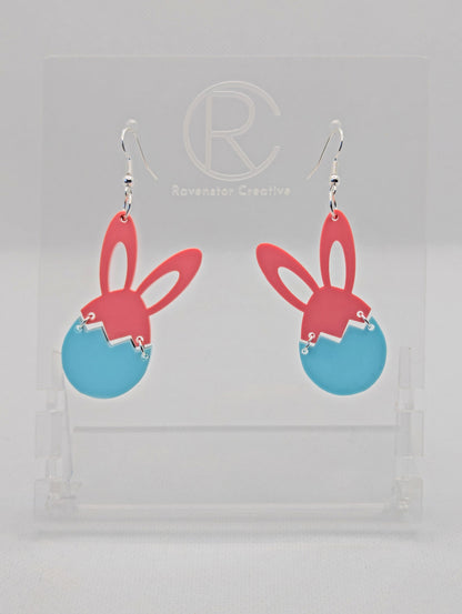 Two part drop earrings with silver ear wire hook fixings. The top part are bunny ears and the bottom is a broken egg joined by jump rings. The colours are Sherbet Pink for the ears and Spearmint for the egg. They are in a clear stand.