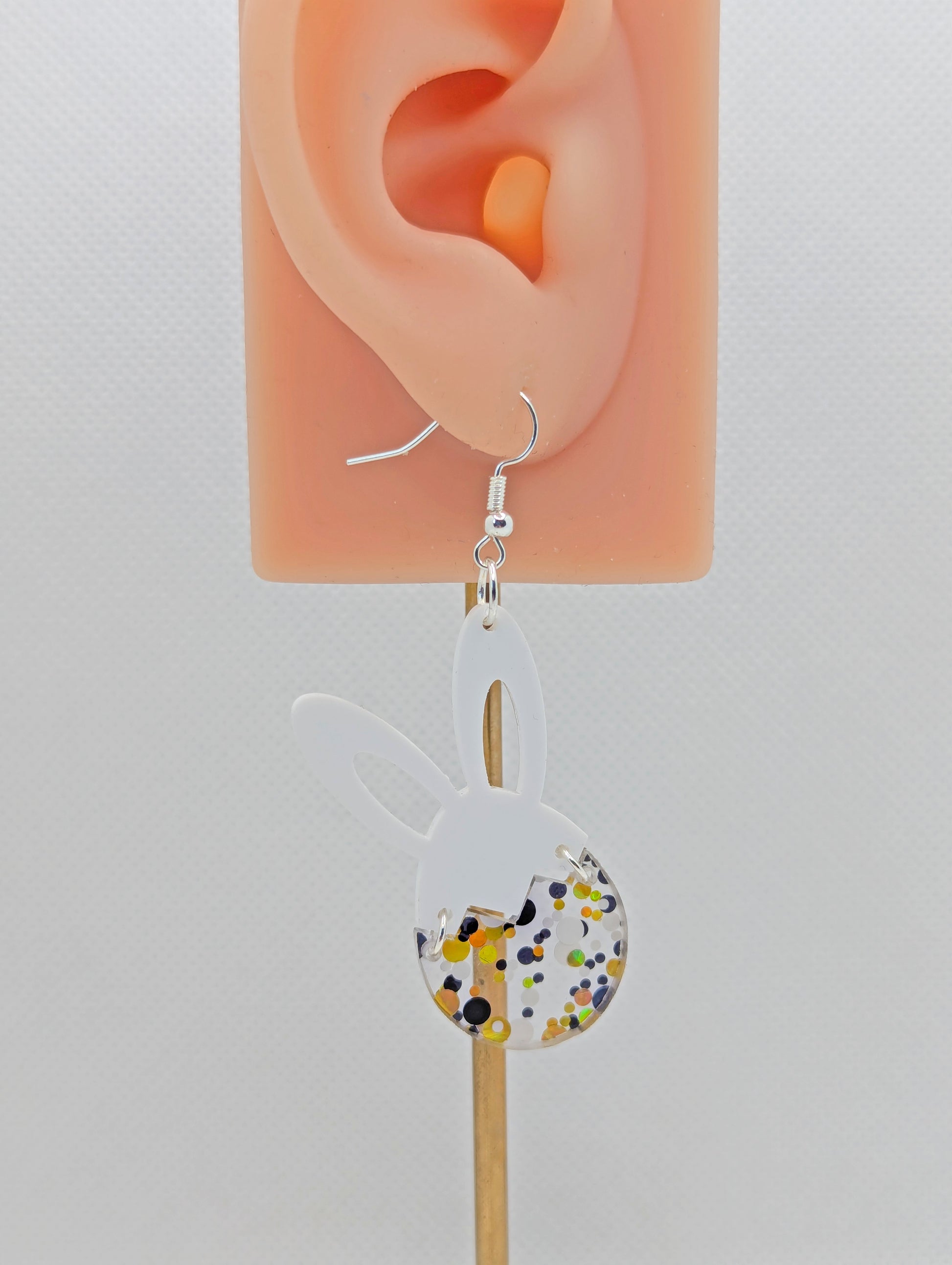 An image of a two part drop earring with a silver ear wire hook. The top part are bunny ears and the bottom is a broken egg joined by jump rings. The colours are White for the ears and Lollipop Glitter for the egg. It is on an ear to show the size.