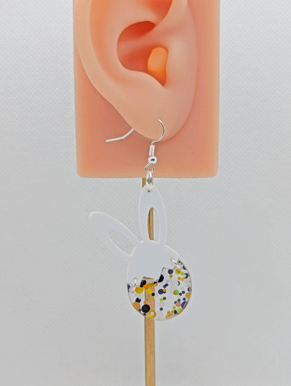 An image of a two part drop earring with a silver ear wire hook. The top part are bunny ears and the bottom is a broken egg joined by jump rings. The colours are White for the ears and Lollipop Glitter for the egg. It is on an ear to show the size.