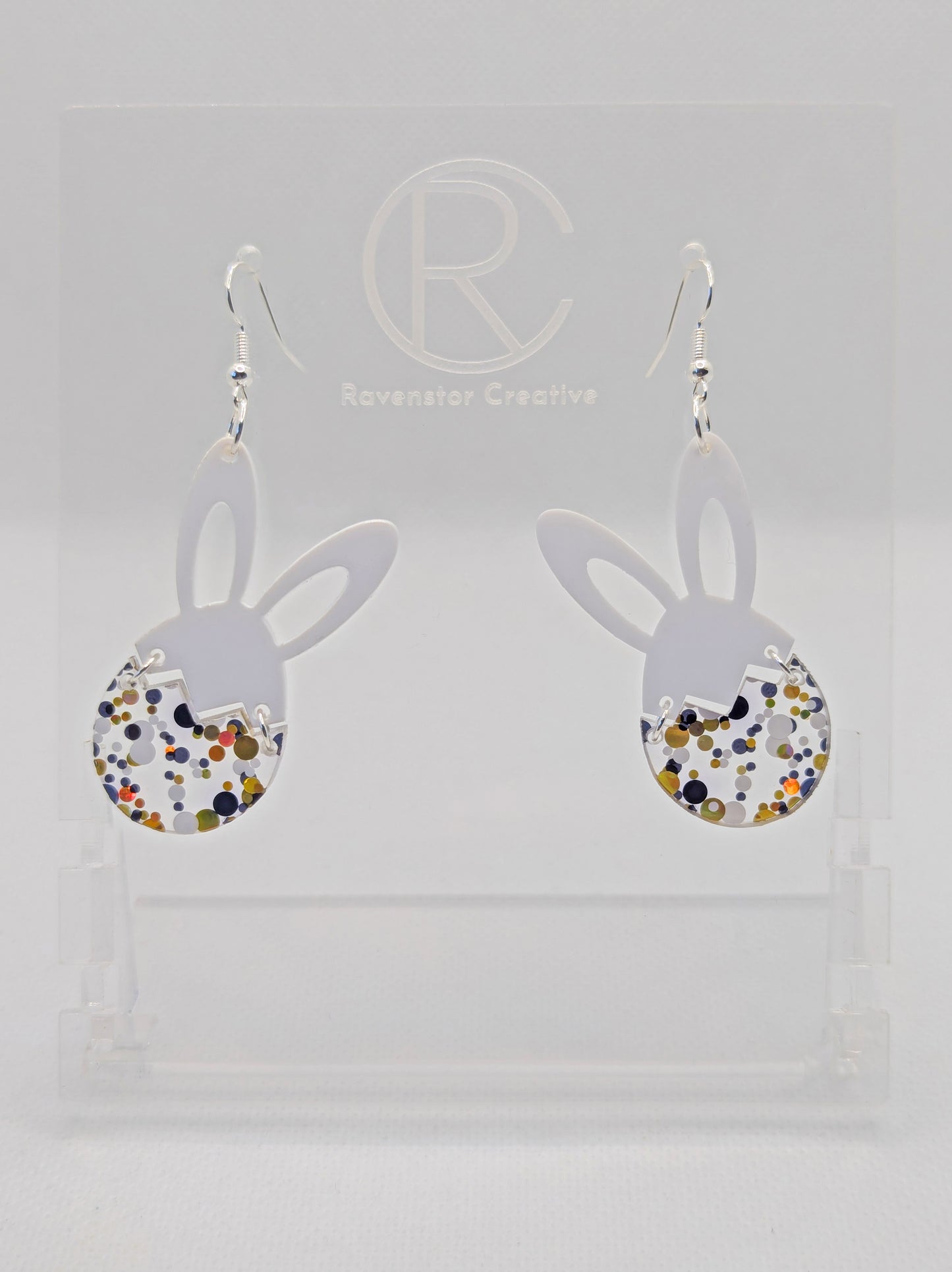 Two part drop earrings with silver ear wire hook fixings. The top part are bunny ears and the bottom is a broken egg joined by jump rings. The colours are White for the ears and Lollipop Glitter for the egg. They are in a clear stand.