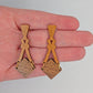 Image of a pair of wooden earrings. The design is a thin elegant top that forks out to support a diamond shape dangling under it. They are against a hand to show the size of three and a half finger widths in length and one wide.