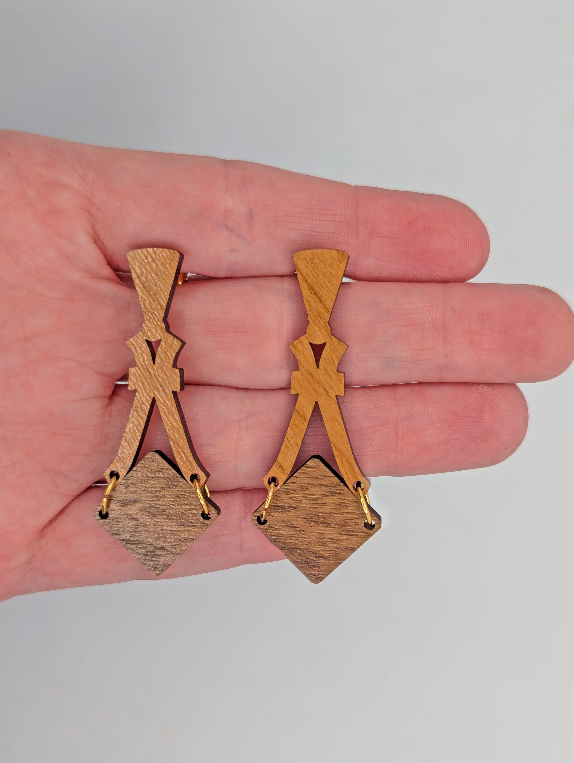 Image of a pair of wooden earrings. The design is a thin elegant top that forks out to support a diamond shape dangling under it. They are against a hand to show the size of three and a half finger widths in length and one wide.