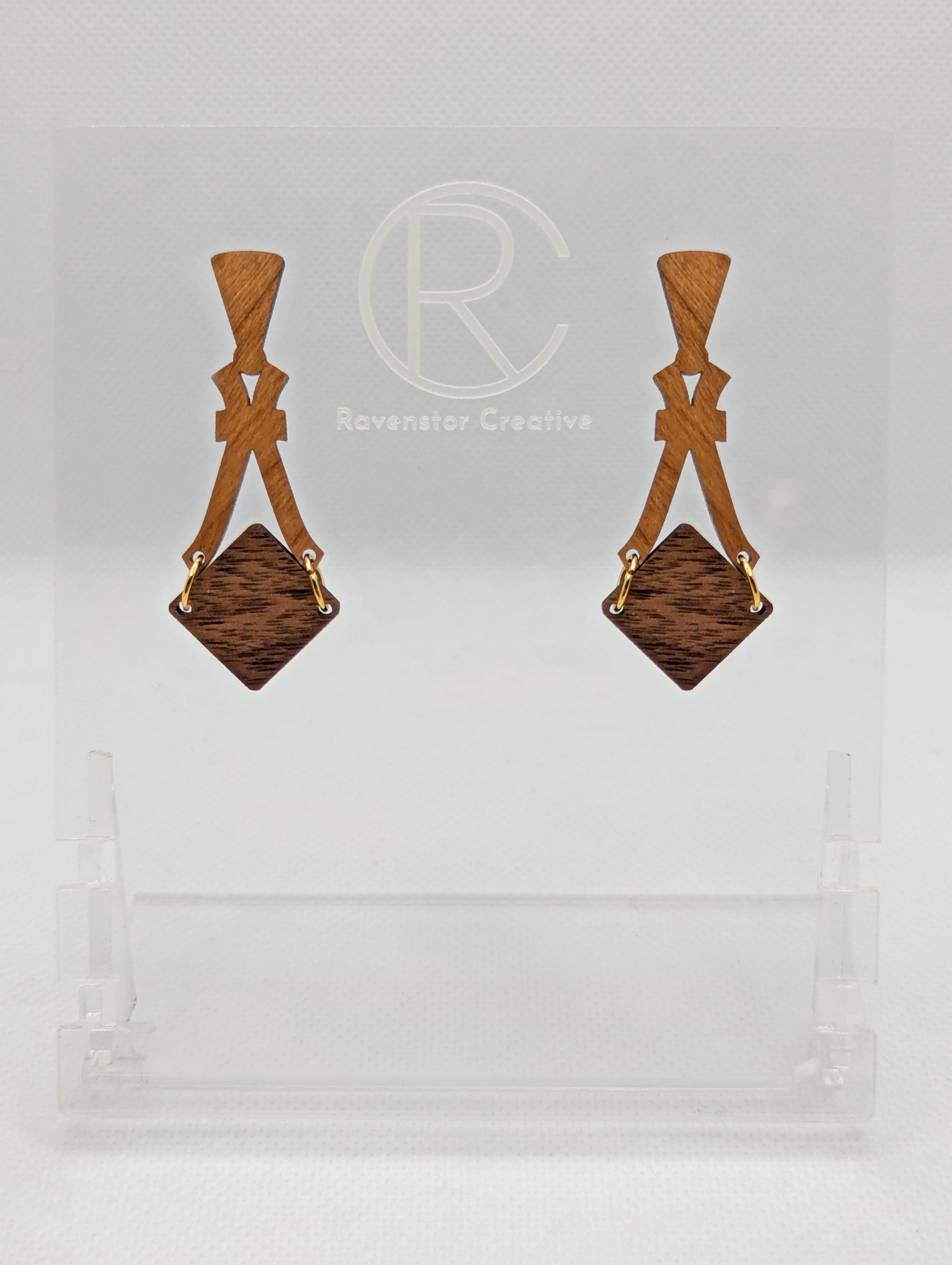 Image of a pair of wooden earrings. The design is a thin elegant top that forks out to support a diamond shape dangling under it. They are Cherry Wood for the top and a Walnut Wood diamond. They are against a white background on a clear stand.